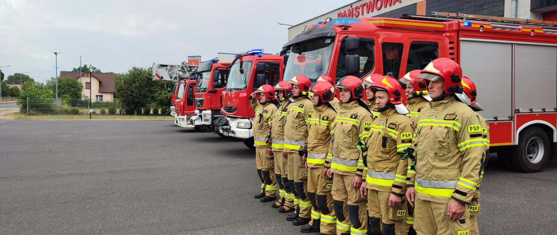 Strażacy stoją w dwuszeregu na tle budynku komendy. Obok stoją pojazdy pożarnicze.
