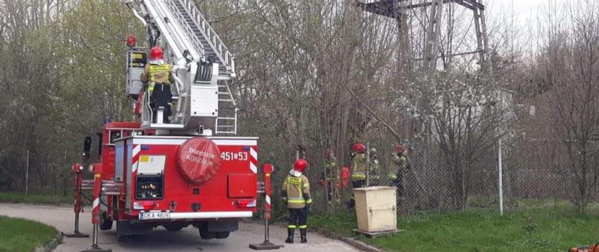 Rozstawiony pojazd strażacki (podnośnik) wysunięte podpory w koszy znajduje się ratownik, kokpit obsługiwany przez siedzącego strażaka w ubraniu specjalnym oraz hełmie. Po prawej stronie widoczny słup energetyczny u jego podstawy za ogrodzeniem znajduje się czterech funkcjonariuszy straży pożarnej