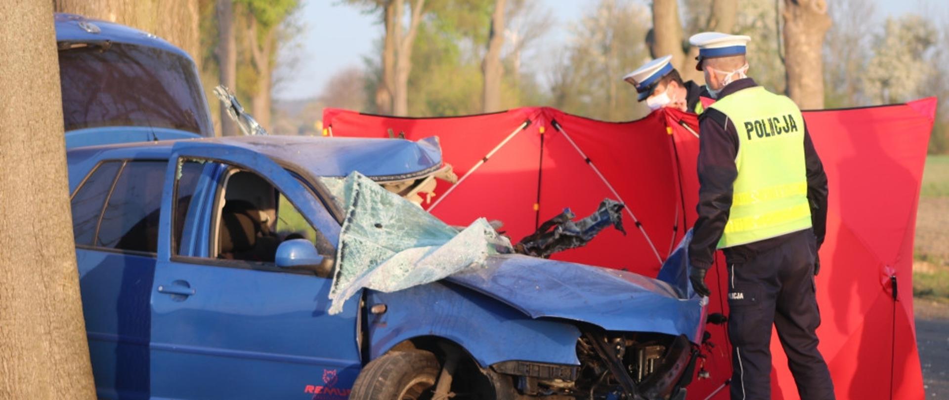 Na zdjęciu znajduje się uszkodzony samochód osobowy po uderzeniu w drzewo oraz dwóch policjantów.