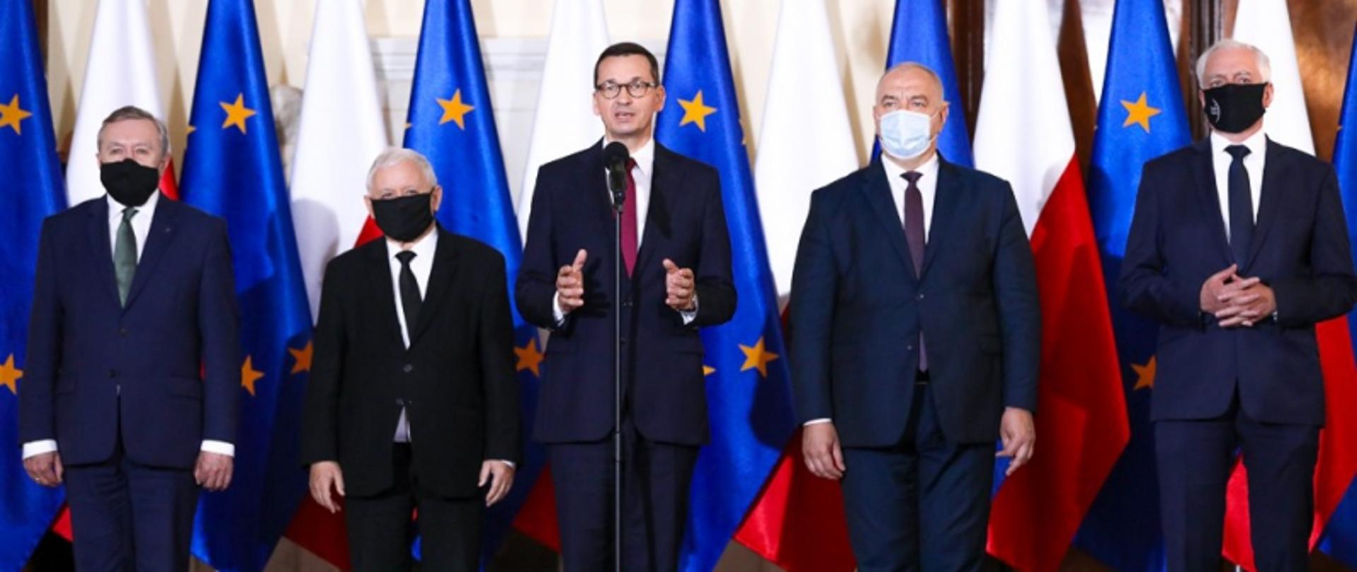 W centralnej części kadru znajduje się premier Polski Mateusz Morawiecki, który przemawia do dziennikarzy zgromadzonych na konferencji prasowej. Obok premiera stoją mężczyźni - nowi ministrowie oraz wicepremierzy. Za mężczyznami 12 flag ustawionych w rzędzie, kolejno flaga Unii Europejskiej oraz flaga RP. 