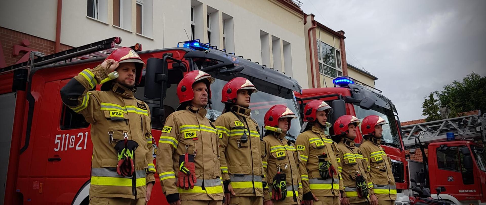 Siedmiu strażaków w umundurowaniu specjalnym oraz hełmach stoi na baczność w szeregu przed samochodami ratowniczo-gaśniczymi. Samochody ustawione są przed garażami od strony ulicy Armii Krajowej, mają włączone sygnały swietlne i dzwiękowe. Dowódca salutuje