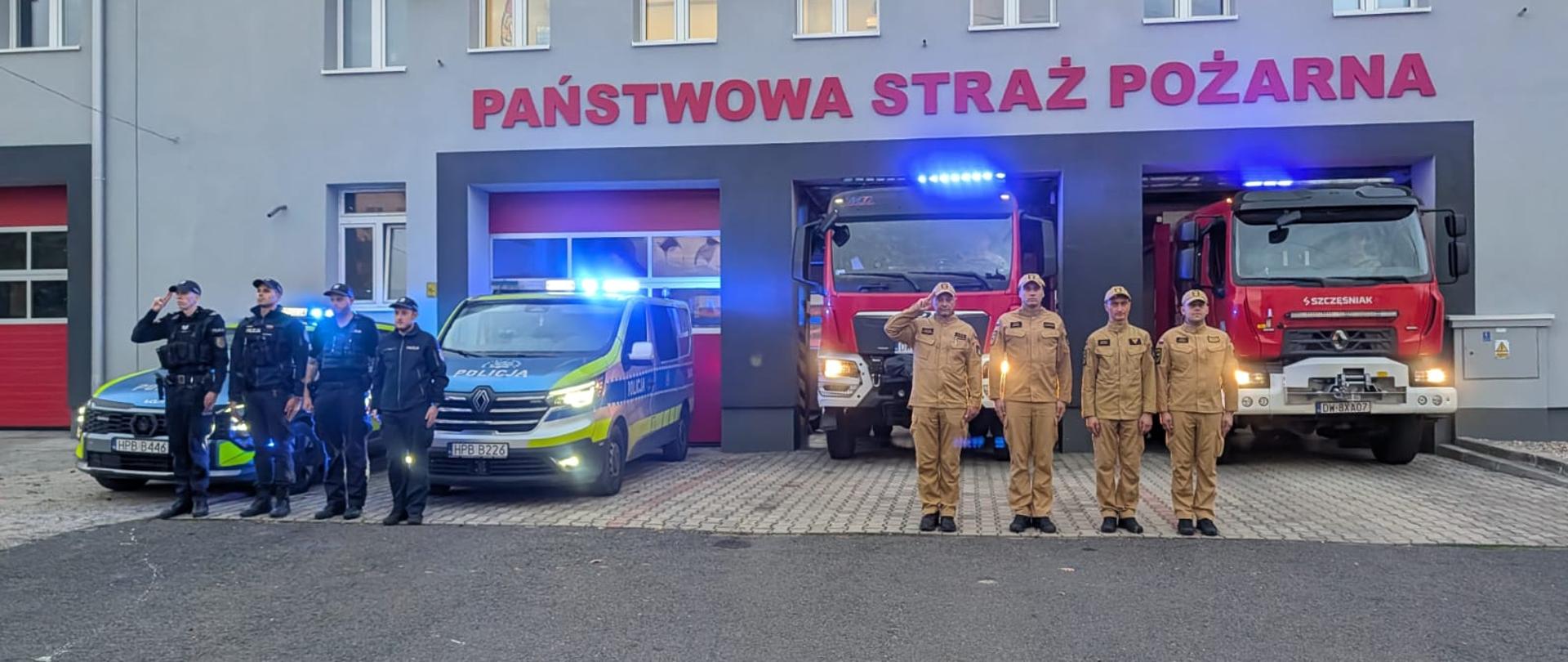 Strażacy w umundurowaniu służbowym i specjalnym stoją przed budynkiem Państwowej Straży Pożarnej, obok samochodów służbowych i pojazdów ratowniczo-gaśniczych z włączonymi niebieskimi sygnałami świetlnymi.