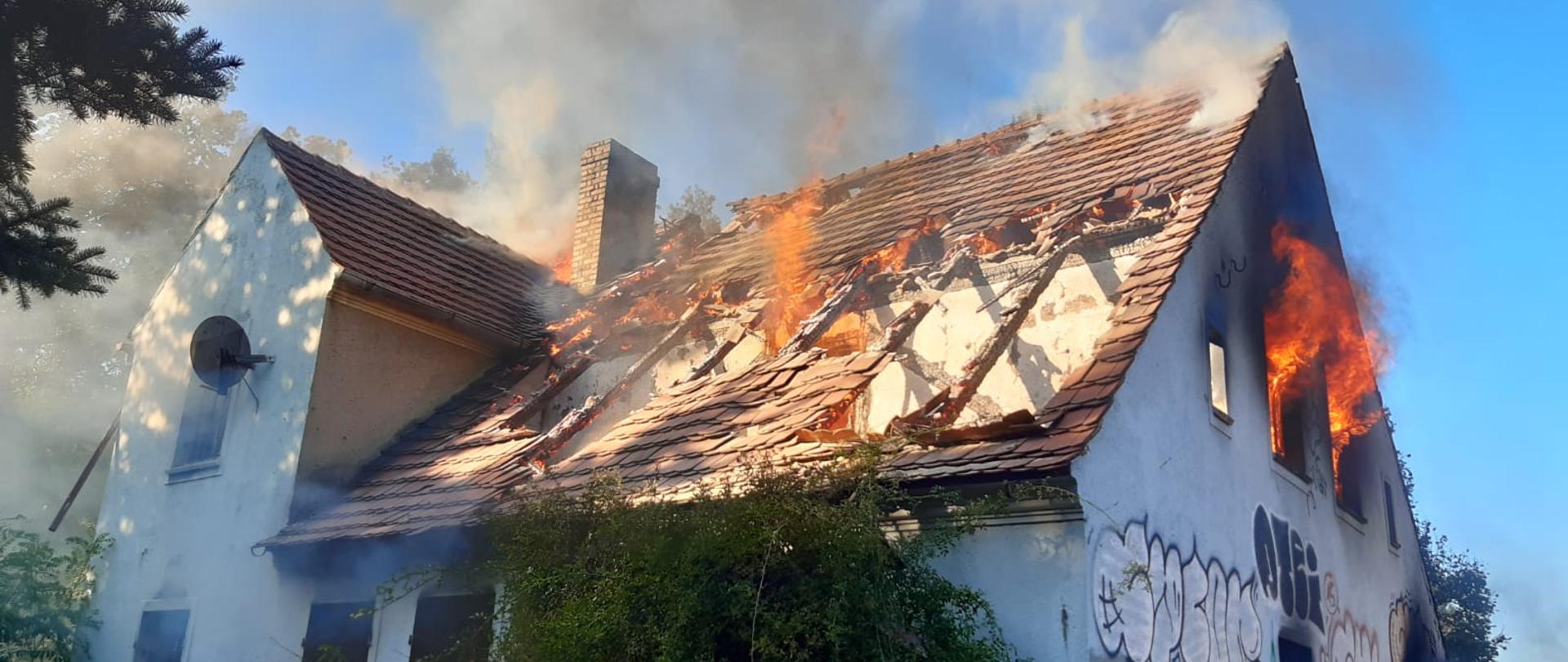 Na zdjęciu znajduje się budynek mieszkalny prawdopodobnie pustostan. Pali się dach i pierwsze piętro. Widać płomienie ognia na dachu i w otworach okiennych.