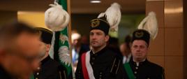 Szereg mężczyzn w czarnych galowych mundurach i górniczych czapkach z białymi piórami, niosą zielony sztandar.