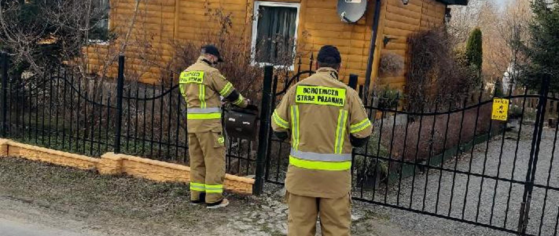 Zdjęcie przedstawia 2 strażaków OSP stojących przed bramą do posesji. Strażacy wkładają ulotkę informacyjną do skrzynki na listy.