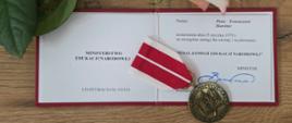 Otwarte etui z legitymacją i Medalem Komisji Edukacji Narodowej leżące na drewnianym blacie. Na dokumentach widoczny jest medal zawieszony na biało-czerwonej wstążce. Nad dokumentami znajduje się jasnoróżowa róża z zielonymi liśćmi. Całość przedstawia uroczyste wręczenie odznaczenia za zasługi dla oświaty i wychowania.