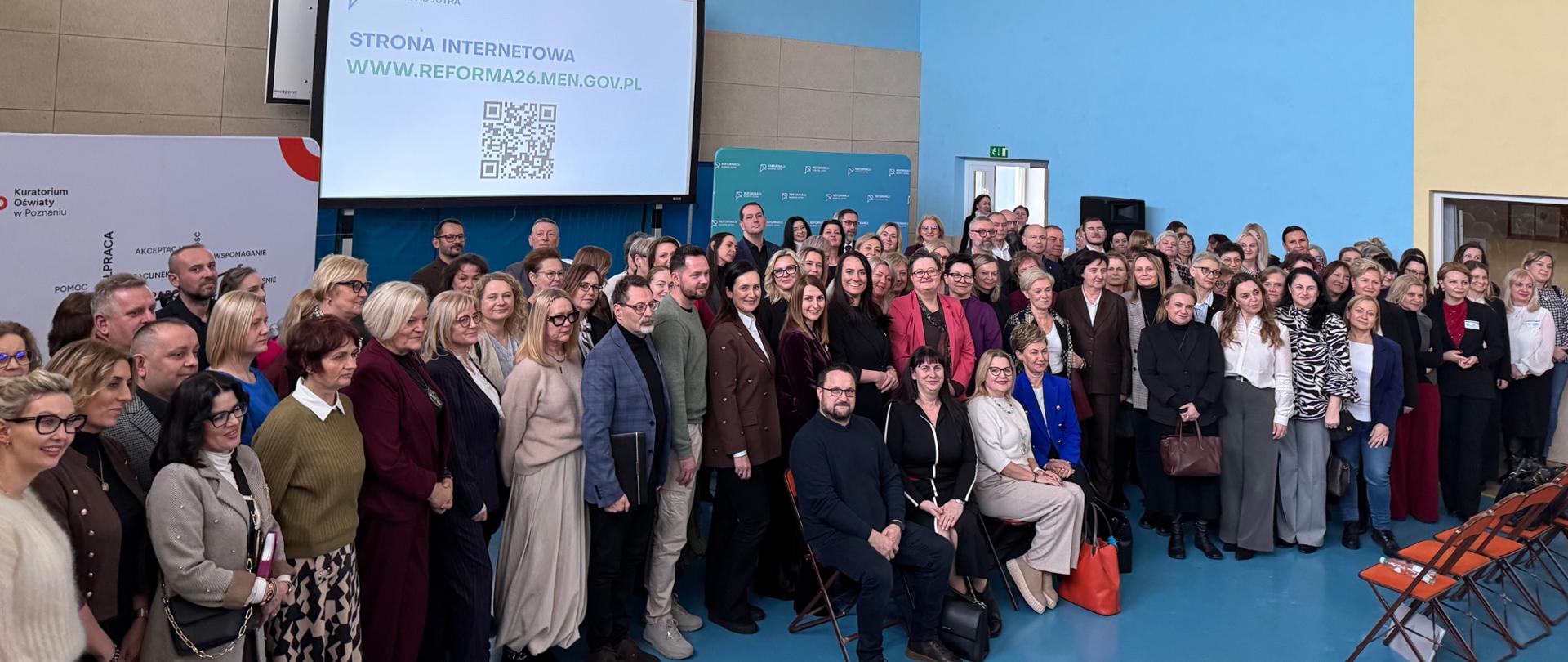 Zdjęcie grupowe kilkudziesięciu osób w hali sportowej. Uczestnicy stoją przed ekranem wyświetlającym adres strony internetowej www.reforma26.men.gov.pl oraz kod QR.