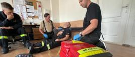 Na zdjęciu na pierwszym planie leży torba medyczna . Na drugim planie jest 4 strażaków. Jeden oparty o łóżko jest poszkodowanym. Po obu jego stronach znajdują się strażacy ćwiczący udzielanie pierwszej pomocy, mają założone rękawiczki nitrylowe. Po lewej stronie na krześle siedzi strażak nadzorujący ćwiczenia.