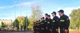 Na zdjęciu widać strażaków zmian służbowych, których wzrok zwrócony się w stronę wciągniętej flagi państwowej. W tle poczet flagowy oddający honor oraz drzewa i niebo częściowo zachmurzone