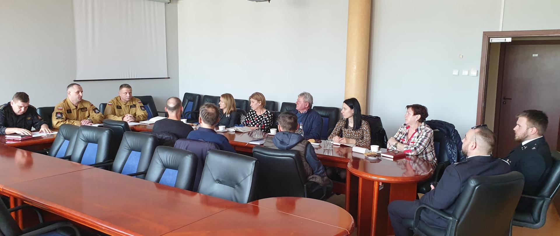 Na zdjęciu widzimy strażaków oraz osoby cywilne siedzące przy stole konferencyjnym. Strażacy ubrani w mundury sztabowe koloru piaskowego oraz czarnego. 