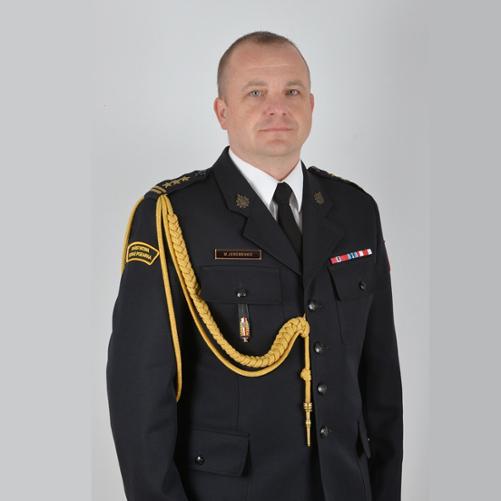 Portret funkcjonariusza Państwowej Straży Pożarnej w mundurze galowym. Mężczyzna stoi na jasnym tle, zwrócony przodem do obiektywu. Ubrany jest w ciemny mundur z białą koszulą i czarnym krawatem. Na lewym ramieniu widoczny jest złoty sznur galowy oraz naszywka „Państwowa Straż Pożarna”. Na piersi znajdują się baretki odznaczeń, odznaka oraz identyfikator z nazwiskiem „M. Jeremenko”. Funkcjonariusz ma krótko przystrzyżone włosy i neutralny wyraz twarzy.