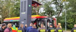 Czerwono – żółty autobus stojący pod stacją ładowania. Z urządzeniem zapoznają się stojący przy nim strażacy i osoby cywilne.