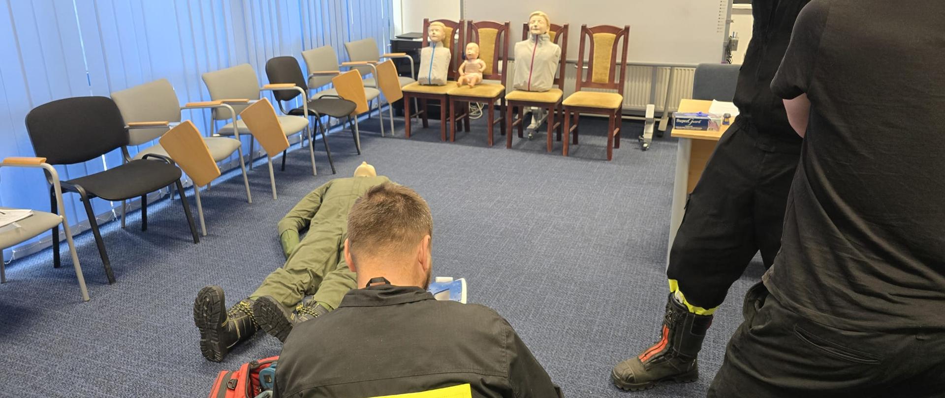 Na zdjęciu widać salę szkoleniową, w której odbywają się ćwiczenia z pierwszej pomocy. W pomieszczeniu znajdują się trzy manekiny szkoleniowe. Dwóch mężczyzn stoi po prawej stronie zdjęcia, obserwując, jak trzeci mężczyzna, ubrany w czarną koszulkę z napisem "STRAŻ" na plecach, klęczy obok manekina leżącego na podłodze w pozycji poszkodowanego. Wszyscy mężczyźni mają na sobie mundury lub strój roboczy. Na podłodze, obok klęczącego, leży czerwona torba, prawdopodobnie torba medyczna.
W głębi sali widać krzesła i ławki, a także białą tablicę, co potwierdza, że jest to sala do ćwiczeń lub szkoleń.
