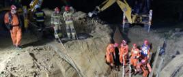 Na zdjęciu w nocy widoczni strażacy w pomarańczowych kombinezonach i czerwonych hełmach i pracujące koparki w trakcie działań ratowniczych mających na celu wydobycie zasypanego ziemią mężczyzny z wykopu.