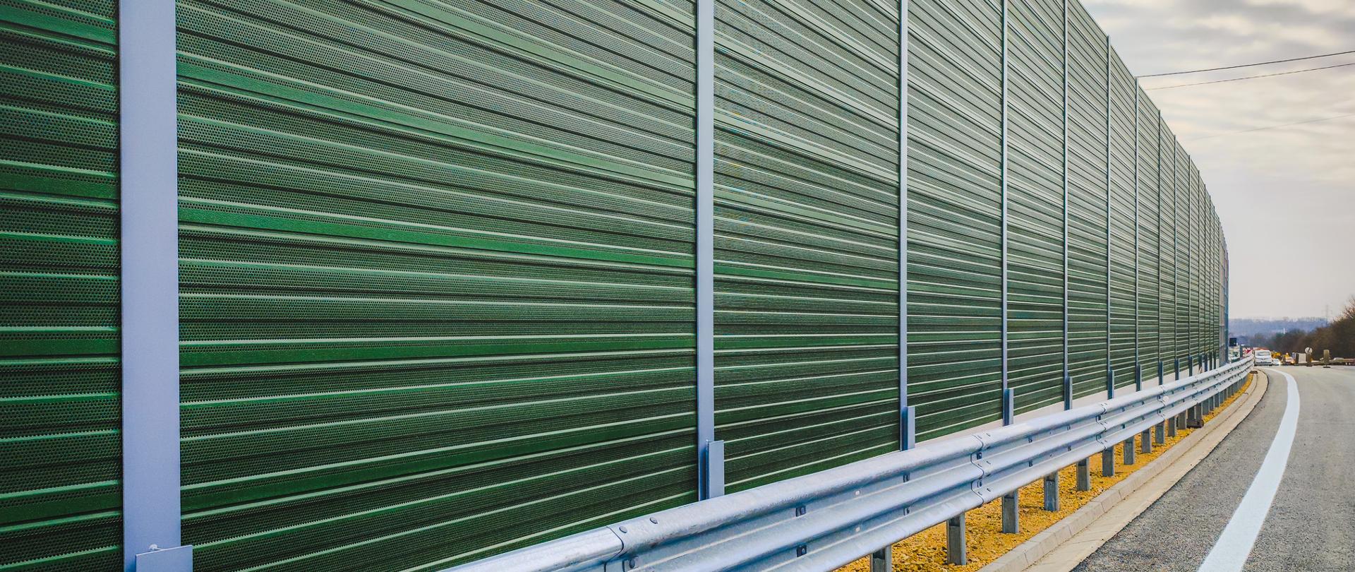 Wysokie, zielone ekrany akustyczne biegnąca wzdłuż zakrętu drogi, z metalową barierą energochłonną u podstawy. Nad drogą widać pochmurne niebo.