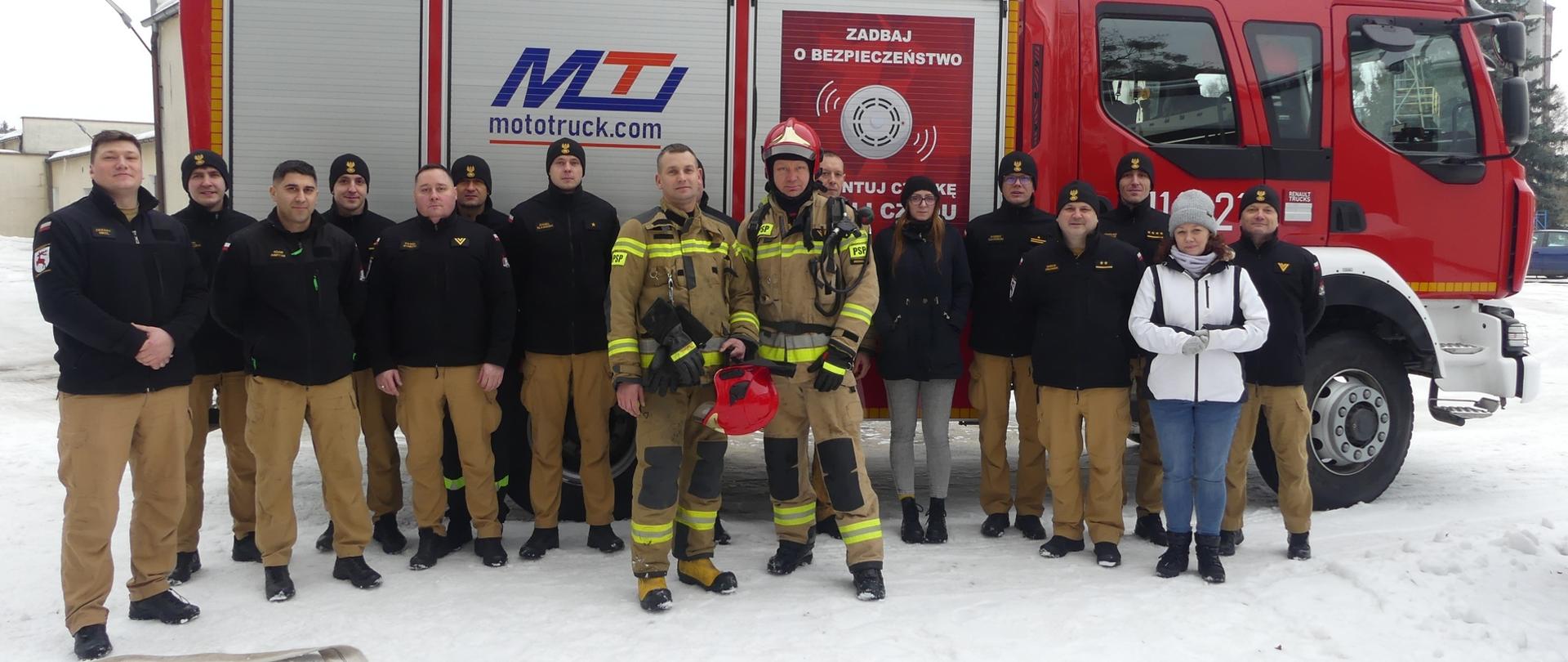 Zdjęcie przedstawia Zbigniewa Bąkowskiego i Karola Magnuszewskiego – w ubraniach specjalnych straży pożarnej – Funkcjonariusze otoczeni przez pracowników Komendy w Brodnicy stoją pozując do pamiątkowego zdjęcia. Z tyłu stoi gaśniczy pojazd JRG w Brodnicy.