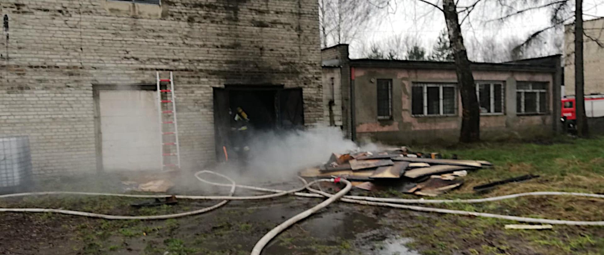 Zdjęcie przedstawia budynek produkcyjny z otwartymi drzwiami. Przed nimi leżą dymiące elementy oraz węże pożarnicze. W drzwiach stoi strażak w ubraniu specjalnym oraz aparacie ochrony układu oddechowego.