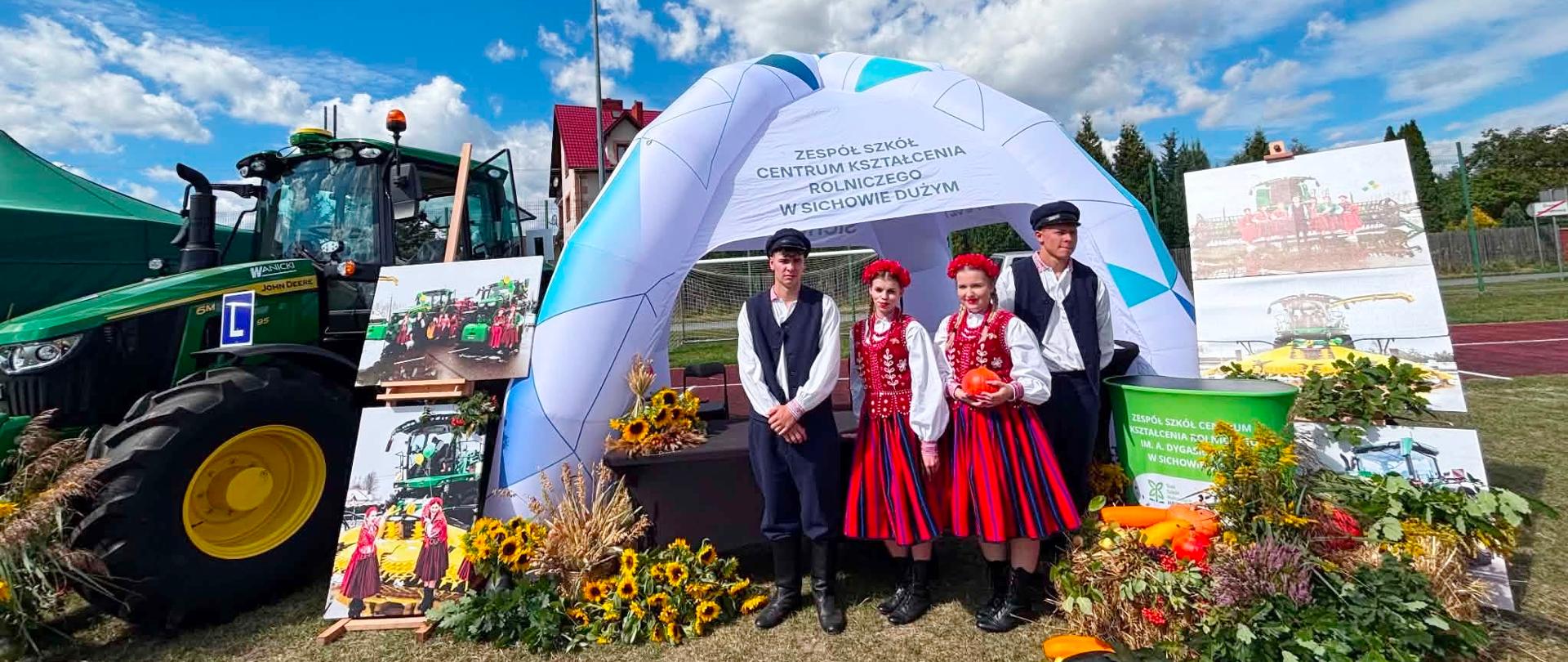 Stoisko wystawiennicze Zespołu Szkół Centrum Kształcenia Rolniczego w Sichowie Dużym podczas dożynek powiatowych w Bogorii. Czworo uczniów w strojach ludowych i historycznych pozuje przed białym, dmuchanym namiotem, obok zielonego ciągnika rolniczego.