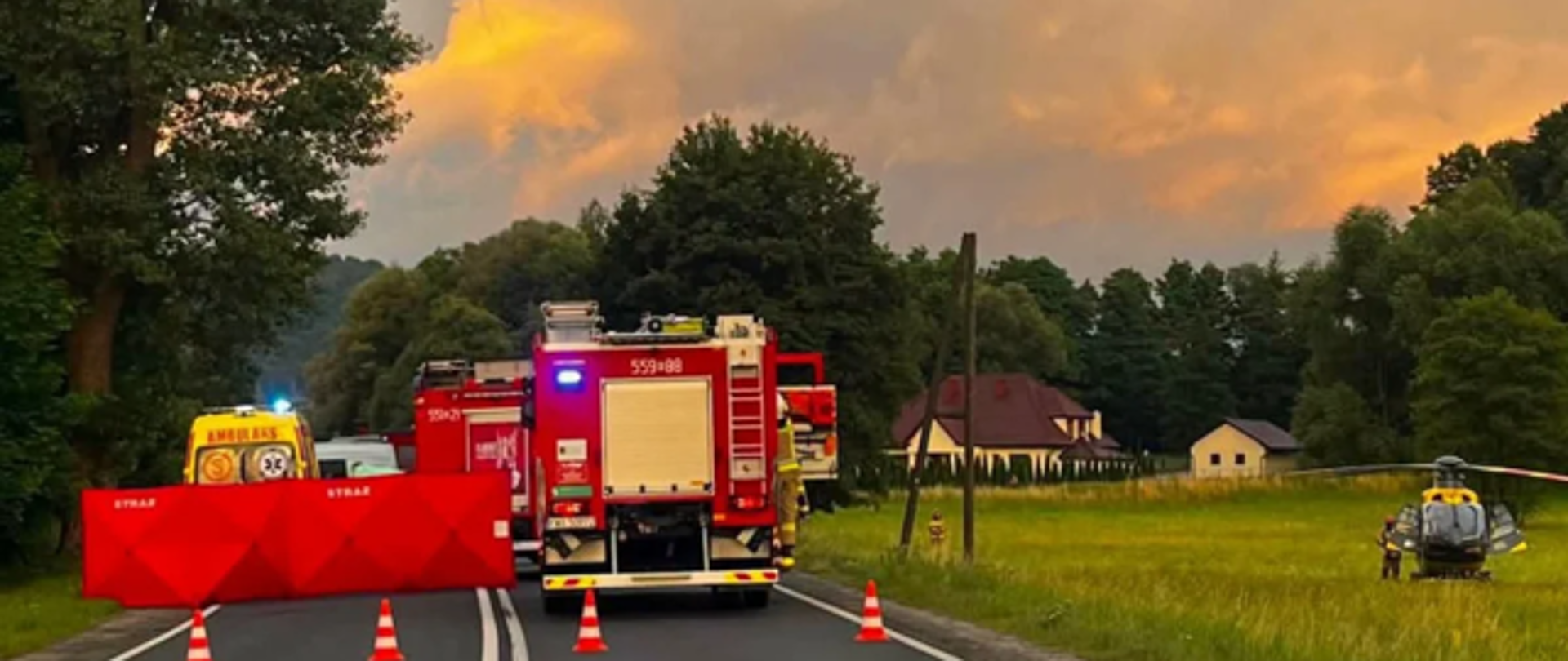 Zdjęcie przedstawia miejsce wypadku. W centrum, na drodze samochody strażackie, z lewej strony karetka. Przed karetką rozłożony czerwony parawan z napisem STRAŻ. Po prawej stronie, na trawie poza drogą stoi śmigłowiec LPR.