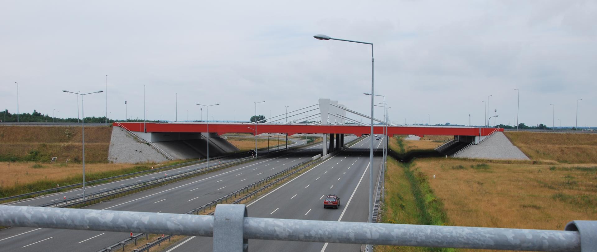 Widok z wiaduktu na obie trzypasowe jezdnie autostrady A2, w tle widoczne wiadukty autostrady A1 z białymi pylonami i naciągami, biegnące górą. Ruch na drodze niewielki.