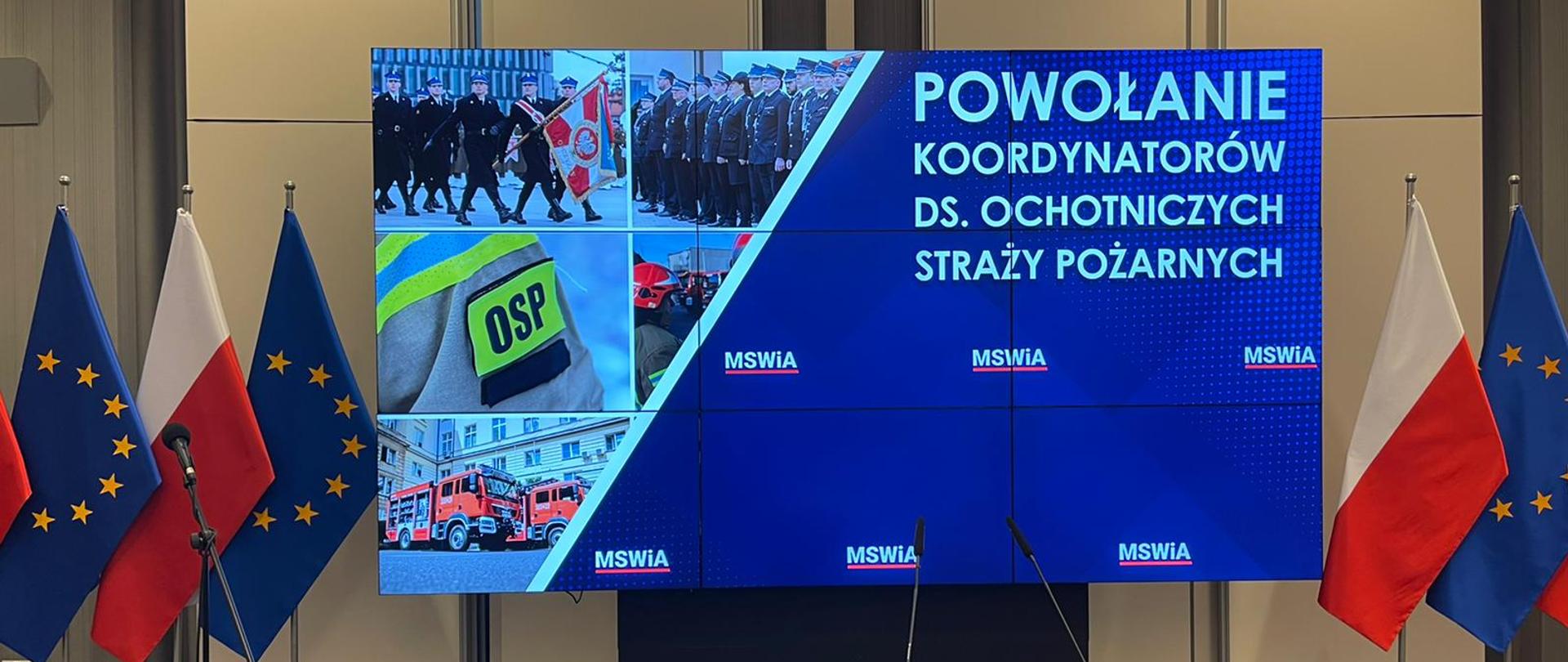 Na telebimie wyświetlany jest napis powołanie koordynatorów do spraw ochotniczych straży pożarnych obok stoją flagi Unii Europejskiej oraz Polski.