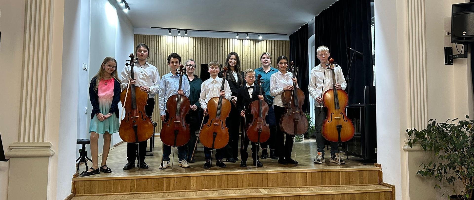 Galeria 10 zdjęć z koncertu uczniów klasy wiolonczeli, który odbył się 19 maja 2025 w auli PSM w Kłodzku. Uczniowie grają na instrumentach w sali koncertowej.