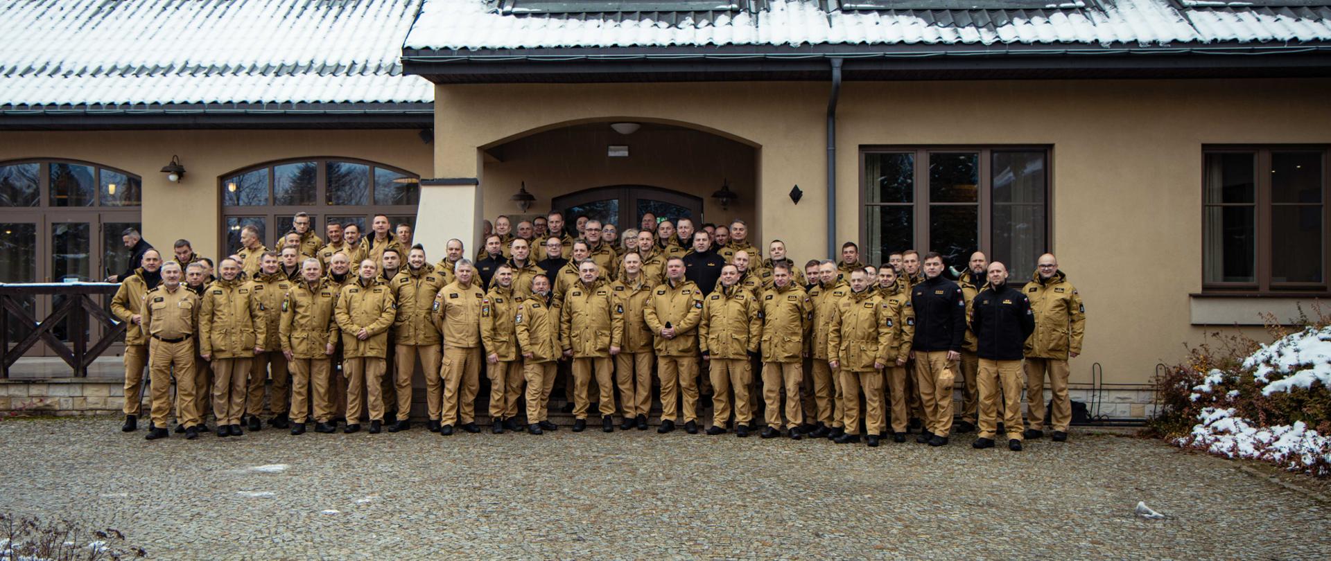 Duża grupa funkcjonariuszy PSP w mundurach wyjściowych pozuje do wspólnego zdjęcia przed budynkiem szkoleniowym; wokół widać zimowy krajobraz z zalegającym śniegiem.