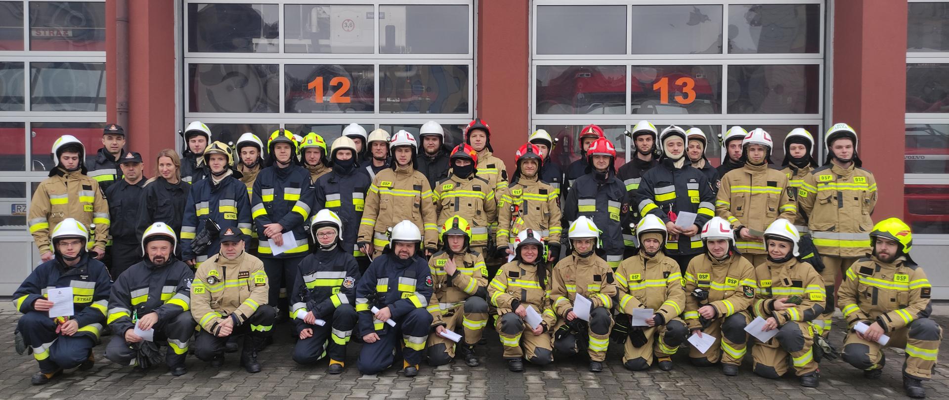 Na zdjęciu widnieją strażacy OSP w umundurowaniu specjalnym. Ustawieni są w 3 rzędach i znajdują się przed garażami jednostki ratowniczo-gaśniczej PSP. W ręku każdy strażak ma papierowe zaświadczenia o ukończeniu szkolenia podstawowego.