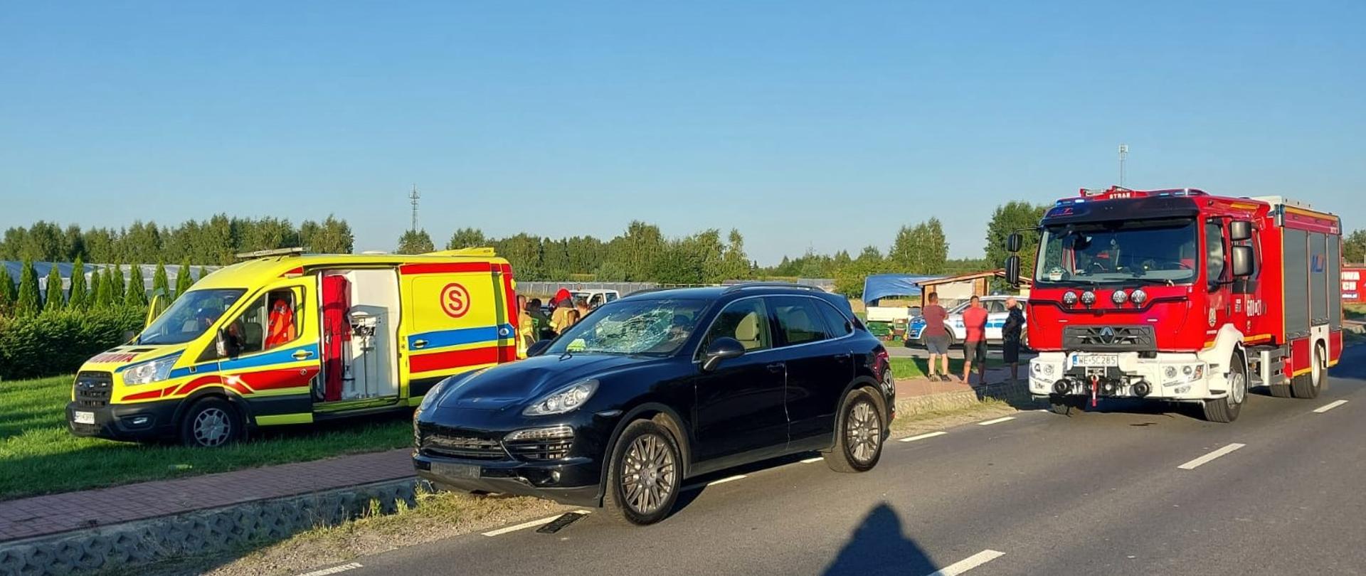 Zdjęcie przedstawia miejsce wypadku drogowego, gdzie na poboczu drogi stoją trzy pojazdy: z lewej żółta karetka pogotowia ratunkowego z niebieskimi i czerwonymi pasami, w środku ciemny samochód osobowy SUV z widocznymi uszkodzeniami przedniej szyby i maski, a po prawej czerwony wóz strażacki. Na drodze widać cień rzucany przez fotografującego. W tle rozciąga się zielony krajobraz z drzewami i linią horyzontu. Pogoda jest słoneczna, a niebo jasne i bezchmurne.