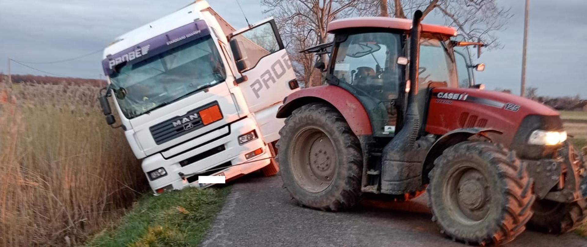 Na zdjęciu biała ciężarówka na zielonym poboczu drogi. Na drodze w poprzek, czerwony traktor.