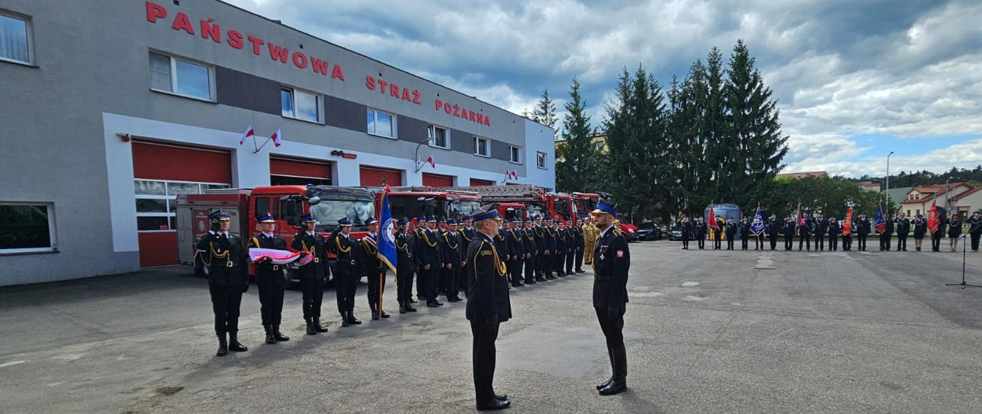 Plac przed budynkiem Państwowej Straży Pożarnej. Zastępca Mazowieckiego Komendanta Wojewódzkiego Państwowej Straży Pożarnej naprzeciw Dowódcy Uroczystości w postawie zasadniczej na środku placu – jeden składa meldunek drugiemu. W tle w rzędzie stoją zaparkowane czerwone wozy strażackie oraz uszykowany w dwuszeregu pododdział strażaków. Po prawej stronie placu widać poczty sztandarowe oraz zaproszonych gości.