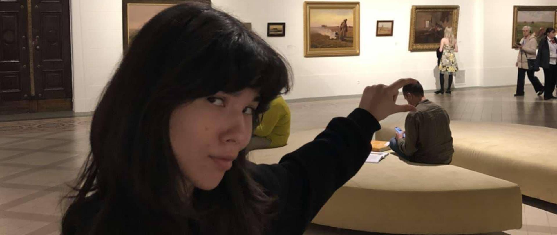 Młoda dziewczyna w dużej przestrzeni wystawienniczej. Wyciąga rękę, pomiędzy jej palcami widać głowę siedzącej opodal osoby