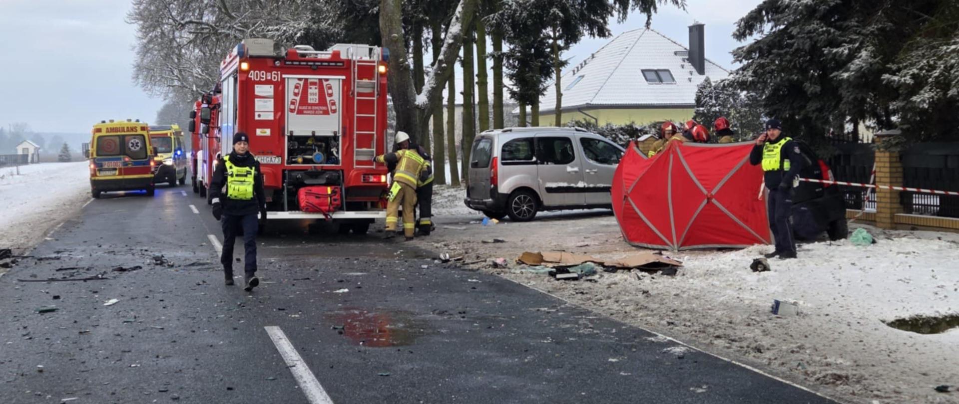 Zdjęcie przedstawia strażaków podczas wypadku drogowego śmiertelnego.
W tle samochody strażackie, samochody uczestniczące w wypadku, ambulanse oraz radiowozy.
