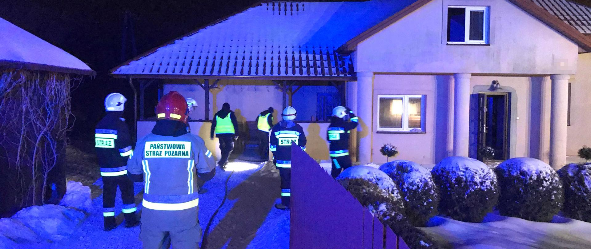 Zdjęcie zrobione w nocy. Zdjęcie przedstawia budynek mieszkalny, w którym pali się pomieszczenie kuchni. Obok domu stoi na ośnieżonym podjeździe pięciu strażaków i dwóch policjantów. 