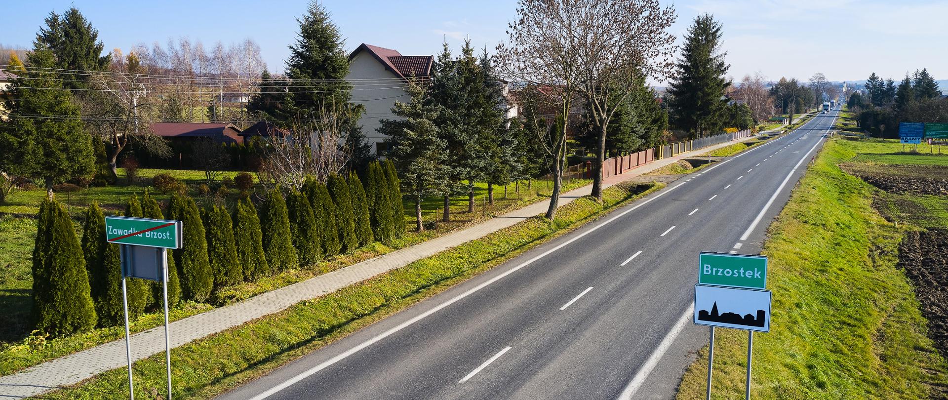 Zdjęcie drogi w letniej scenerii. Świeci słońce. Po prawej stronie znak drogowy z napisem Brzostek. 