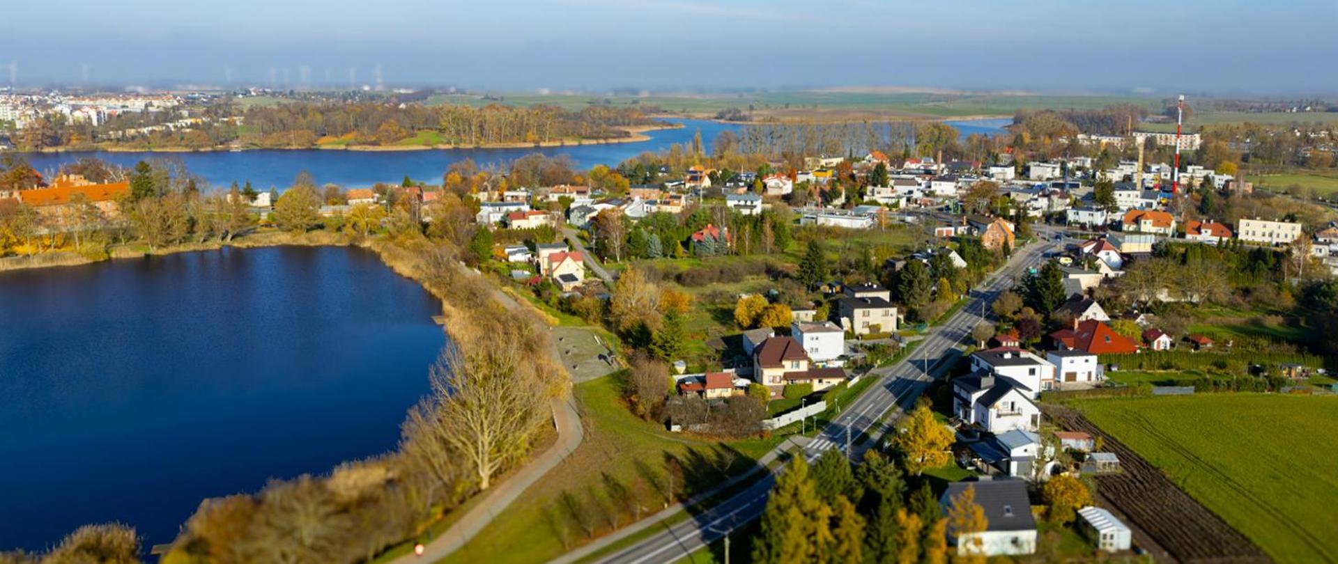 Widok z lotu ptaka na małe miasto nad rzeką lub jeziorem; wzdłuż brzegu biegnie ścieżka, po prawej stronie widoczne domy jednorodzinne i droga, a w tle pola oraz linia drzew.
