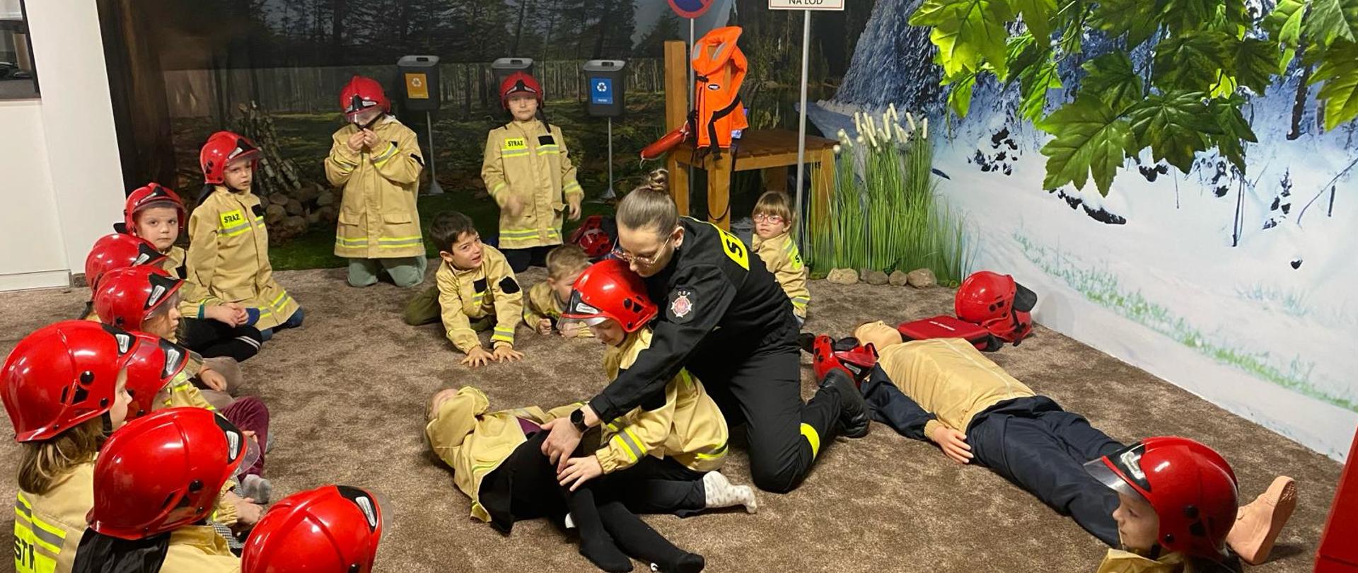 Grupa dzieci podczas zajęć na sali edukacyjnej Ognik. Dzieci siedzą ubrane w specjalnie przygotowane dla nich mundury strażackie po środku widoczne dzieci ćwiczęce wraz z ubraną w czarny mundur funkcjonariuszką pozycję bezpieczną 