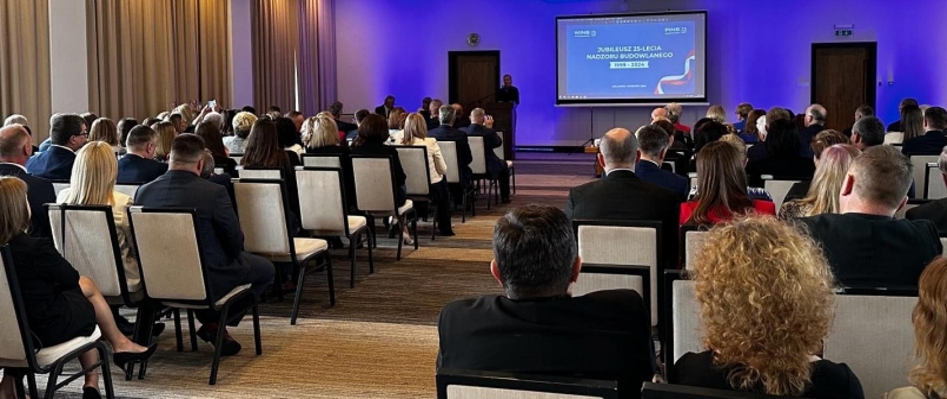 Dorota Cabańska, Główny Inspektor Nadzoru Budowlanego (GINB), podczas uroczystości jubileuszu 25-lecia terenowych organów nadzoru budowlanego na Podkarpaciu. Zdjęcia przedstawiają momenty wręczania odznaczeń i gratulacji wyróżnionym osobom, wspólne zdjęcie grupowe uczestników z odznaczeniami, przebieg konferencji w sali z prezentacją multimedialną, wystąpienia Doroty Cabańskiej oraz ogólną atmosferę wydarzenia w eleganckiej, nowoczesnej sali konferencyjnej.