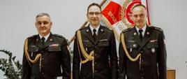 Trzech oficerów PSP w galowych mundurach pozuje do wspólnego zdjęcia na tle sztandaru. Dwóch z nich ma przypięte sznury galowe, wszyscy prezentują odznaczenia i dystynkcje służbowe.