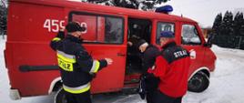 Na zdjęciu widać dwóch strażaków, którzy pomagają pacjentowi wejść do samochodu strażackiego. wszystko odbywa się w scenerii pięknej zimy.