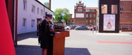 Na zdjęciu widać strażaczkę w galowym mundurze stojącą przy drewnianym podium podczas uroczystości z okazji Dnia Strażaka. W tle stoją szeregi strażaków oraz zabytkowe ceglane budynki i wieża ćwiczeniowa straży pożarnej z banerem wydarzenia. Ceremonia odbywa się na placu jednostki w słoneczny dzień.