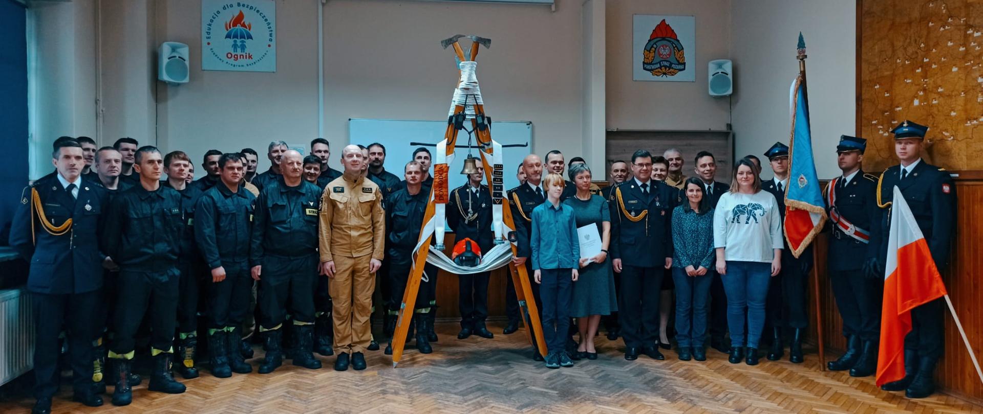 Widok z przodu. Zdjęcie grupowe w świetlicy JRG Wadowice. Pośrodku drabina symbolizująca przebieg służby odchodzącego w stan pozasłużbowy strażaka. 