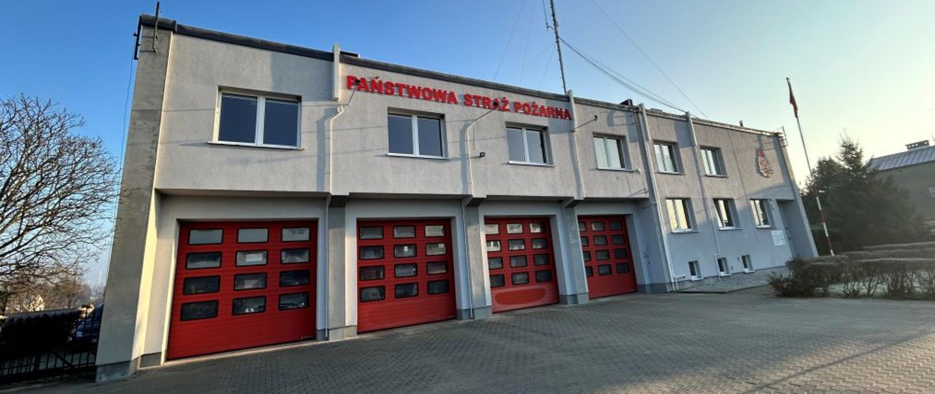 Na zdjęciu znajduje się budynek Państwowej Straży Pożarnej o jasnej elewacji i czerwonych bramach garażowych. Nad garażami widnieje czerwony napis “PAŃSTWOWA STRAŻ POŻARNA”. Budynek ma dwa piętra i kilka okien, a na dachu zamontowane są anteny. Na prawo od budynku widać maszt z powiewającą polską flagą. Przed strażnicą znajduje się brukowany plac, prawdopodobnie służący do manewrowania pojazdami.