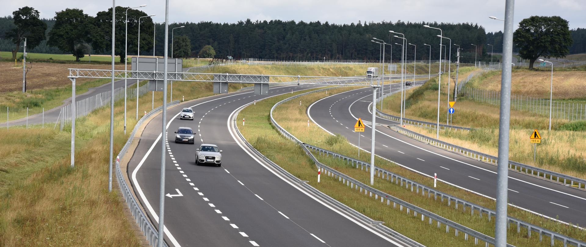 Dwujezdniowa droga wśród łąk. Na horyzoncie widać las. Na drodze znajduje się kilka samochodów.