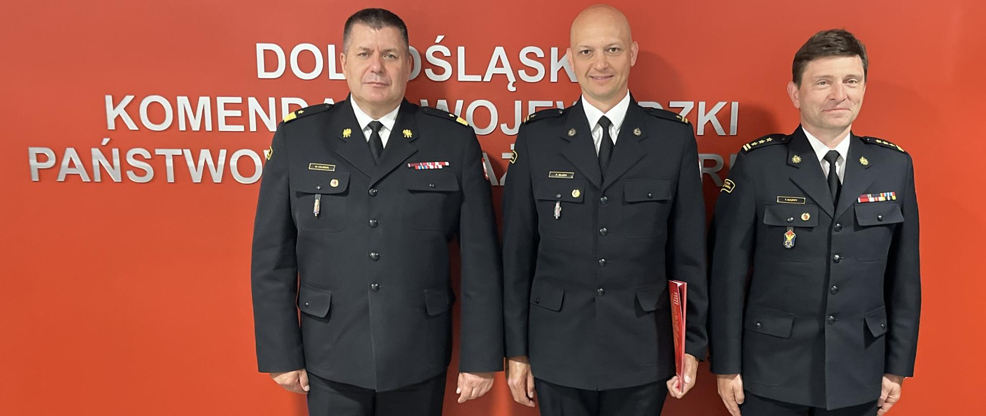 Trzech strażaków w ciemnogranatowych mundurach wyjściowych, białych koszulach i czarnych krawatach, stojących obok siebie. Mężczyźni noszą oficjalne odznaczenia i stopnie służbowe na naramiennikach, a strażak w środku trzyma pod ramieniem czerwoną teczkę. Stoją oni na tle gładkiej, jaskrawoczerwonej ściany.