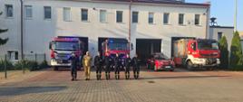 Zdjęcie przedstawia strażaków stojących przed strażnicą podczas oddania hołdu zmarłemu druhowi. Z tyłu stoją samochody strażackie na włączonych sygnałach świetlnych. 