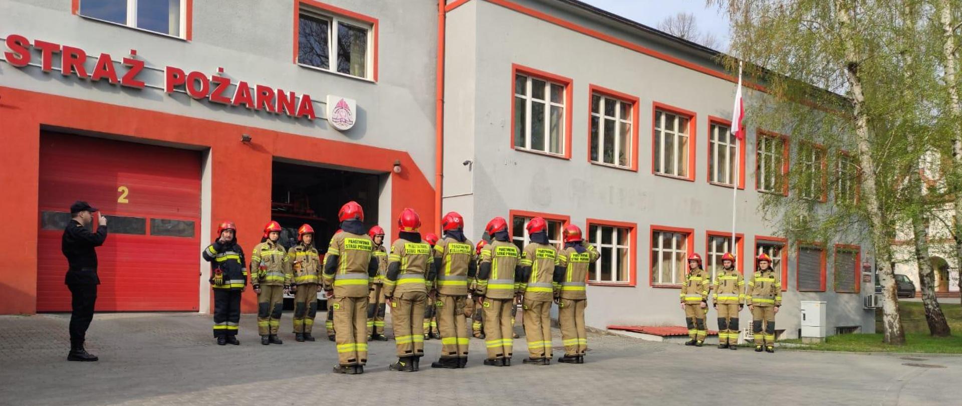 Na zdjęciu strażacy dwóch zmian służbowych w szyku. Wszyscy w ubraniach specjalnych. Przy maszcie z flagą państwową trzech strażaków. Przed zmianami Dowódca JRG przeprowadzający uroczystą zmianę służby z podniesieniem flagi państwowej na maszt.