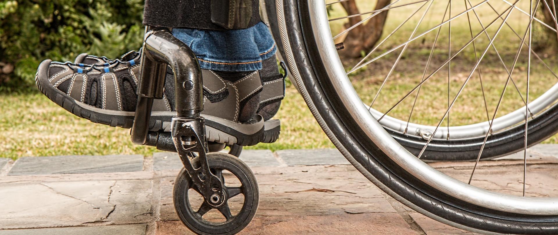 Na zdjęciu widoczna jest dolna część wózka inwalidzkiego. Na wózku siedzi mężczyzna - widoczne są męskie buty oraz długie spodnie. W dalszej części zdjęcia zielone rośliny. 