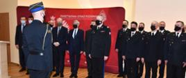 Strażak w mundurze wyjściowym melduje nadbrygadierowi za nim inni strażacy w mundurach oraz osoby w garniturach