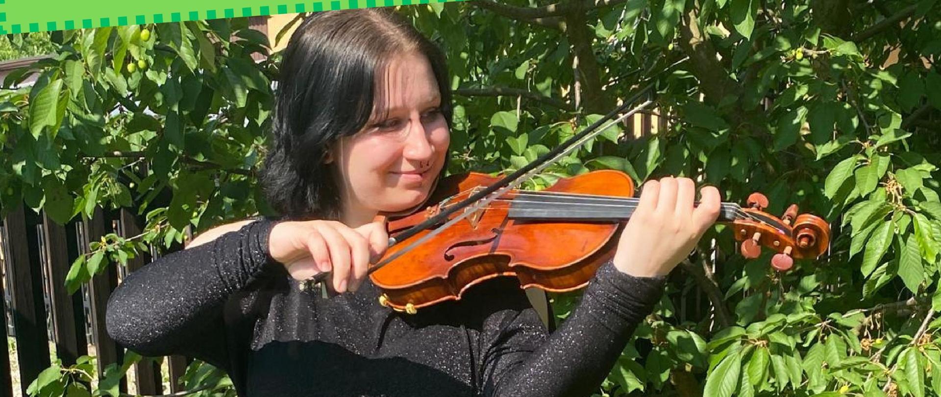 Zdjęcie całej postaci młodej dziewczyny w czerni ze skrzypcami ułożonymi w pozycji grającej. W tle trawa i drzewa. U góry litery z informacją o recitalu. Na dole niebieskie logo szkoły.